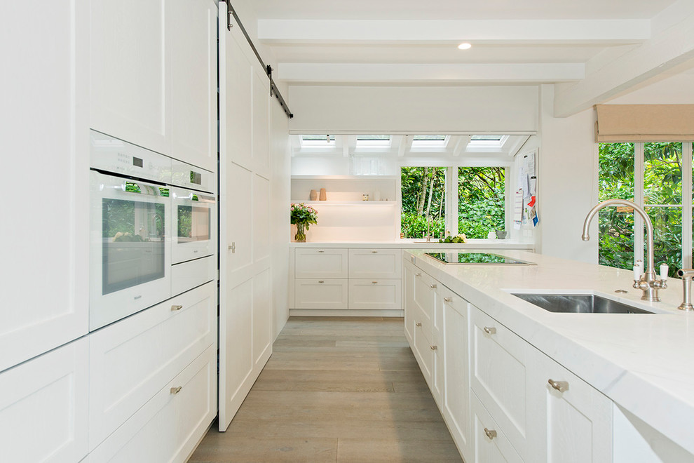 Inspiration for a large traditional l-shaped open plan kitchen in Auckland with an undermount sink, shaker cabinets, white cabinets, quartz benchtops, white splashback, white appliances, light hardwood floors, with island and beige floor.