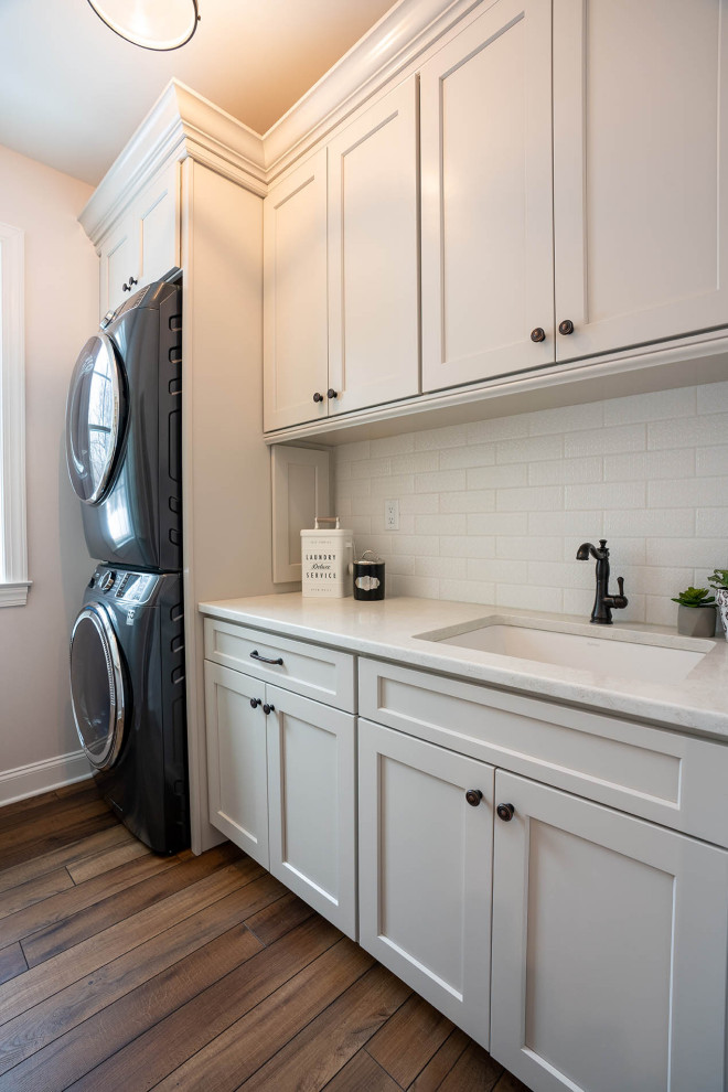 Photo of a large contemporary l-shaped laundry room in Philadelphia with an undermount sink, recessed-panel cabinets, white cabinets, quartz benchtops, grey splashback, ceramic splashback, vinyl floors, brown floor and white benchtop.