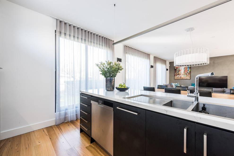 This is an example of a large modern galley open plan kitchen in Melbourne with a double-bowl sink, flat-panel cabinets, black cabinets, quartz benchtops, white splashback, ceramic splashback, black appliances, light hardwood floors, with island, brown floor and white benchtop.