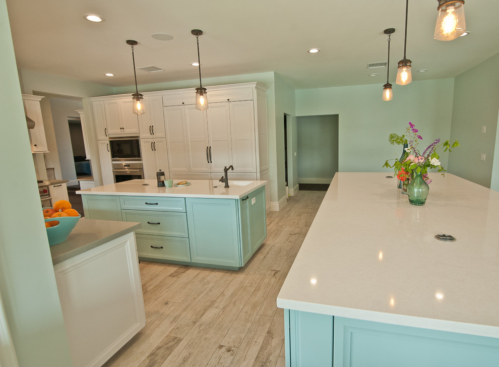 This is an example of an expansive beach style u-shaped open plan kitchen in Orange County with a farmhouse sink, raised-panel cabinets, white cabinets, quartz benchtops, blue splashback, ceramic splashback, stainless steel appliances, ceramic floors and multiple islands.