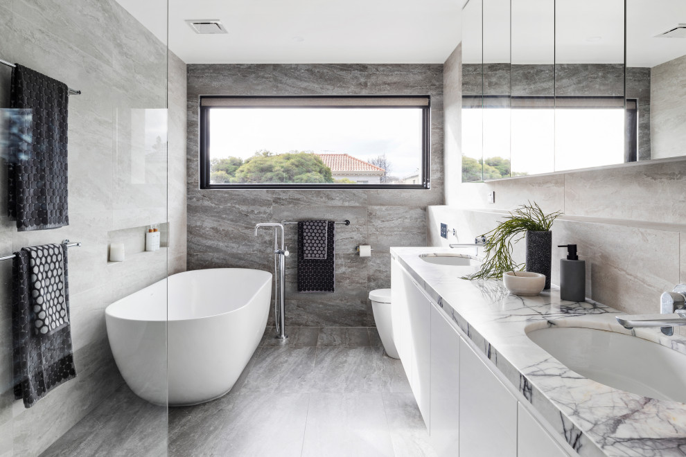 Large modern master bathroom in Melbourne with white cabinets, a freestanding tub, a one-piece toilet, gray tile, an undermount sink, grey floor, an open shower, porcelain tile, grey walls, porcelain floors, marble benchtops, an open shower, white benchtops and flat-panel cabinets.
