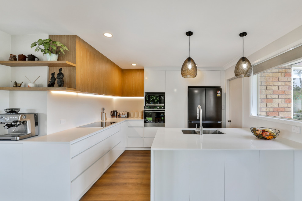 This is an example of a medium sized contemporary l-shaped kitchen/diner in Auckland with a double-bowl sink, flat-panel cabinets, white cabinets, engineered stone countertops, white splashback, engineered quartz splashback, stainless steel appliances, vinyl flooring, a breakfast bar, brown floors and white worktops.