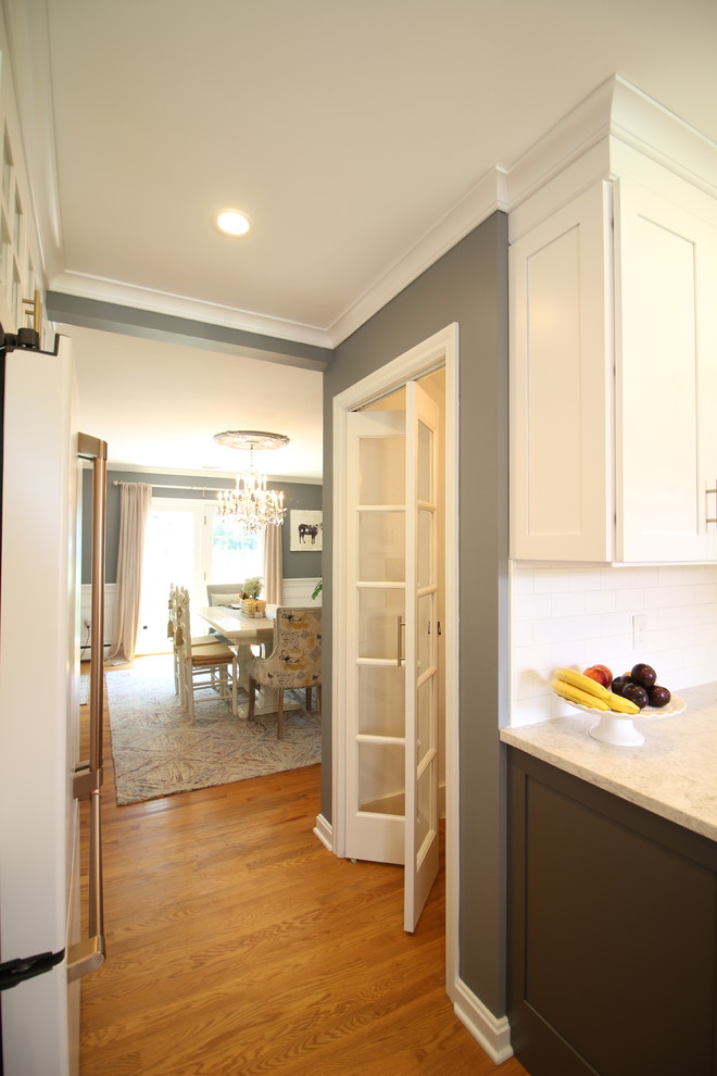 Example of a mid-sized transitional u-shaped medium tone wood floor and brown floor enclosed kitchen design with a farmhouse sink, shaker cabinets, white cabinets, quartz countertops, white backsplash, subway tile backsplash, white appliances, no island and beige countertops
