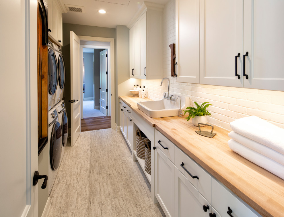 Mid-sized transitional single-wall utility room in Minneapolis with flat-panel cabinets, white cabinets, wood benchtops, white walls, laminate floors, a stacked washer and dryer, grey floor, a farmhouse sink and white benchtop.