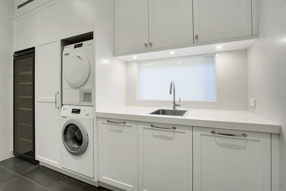 Mid-sized traditional single-wall utility room in Sydney with a single-bowl sink, shaker cabinets, white cabinets, quartz benchtops, white walls, slate floors, a stacked washer and dryer and grey floor.