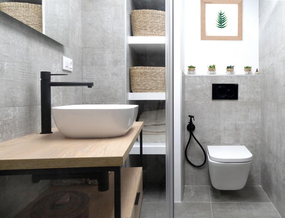 Modern inredning av ett mellanstort brun brunt badrum med dusch, med möbel-liknande, skåp i ljust trä, en kantlös dusch, en toalettstol med hel cisternkåpa, grå kakel, vita väggar, ett fristående handfat, träbänkskiva, grått golv och dusch med gångjärnsdörr
