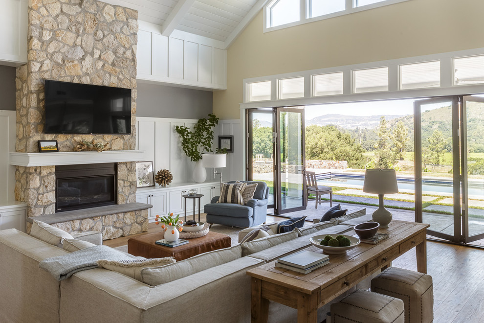 Large country open concept family room in San Francisco with yellow walls, light hardwood floors, a standard fireplace, a stone fireplace surround, a wall-mounted tv and beige floor.