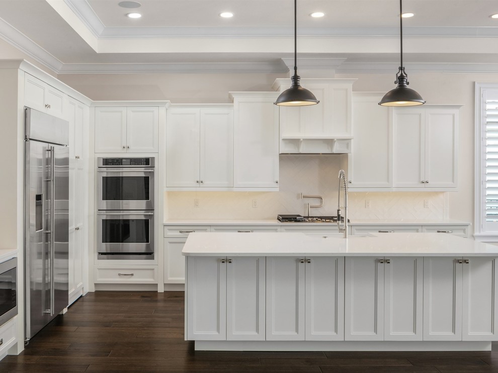 Mid-sized transitional l-shaped open plan kitchen in Orlando with recessed-panel cabinets, white cabinets, white splashback, stainless steel appliances, dark hardwood floors, with island, brown floor, white benchtop and a farmhouse sink.