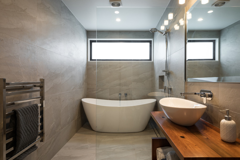 Mid-sized contemporary master wet room bathroom in Other with open cabinets, medium wood cabinets, a freestanding tub, a wall-mount toilet, ceramic tile, grey walls, ceramic floors, a vessel sink, wood benchtops, grey floor, an open shower, beige tile and brown benchtops.