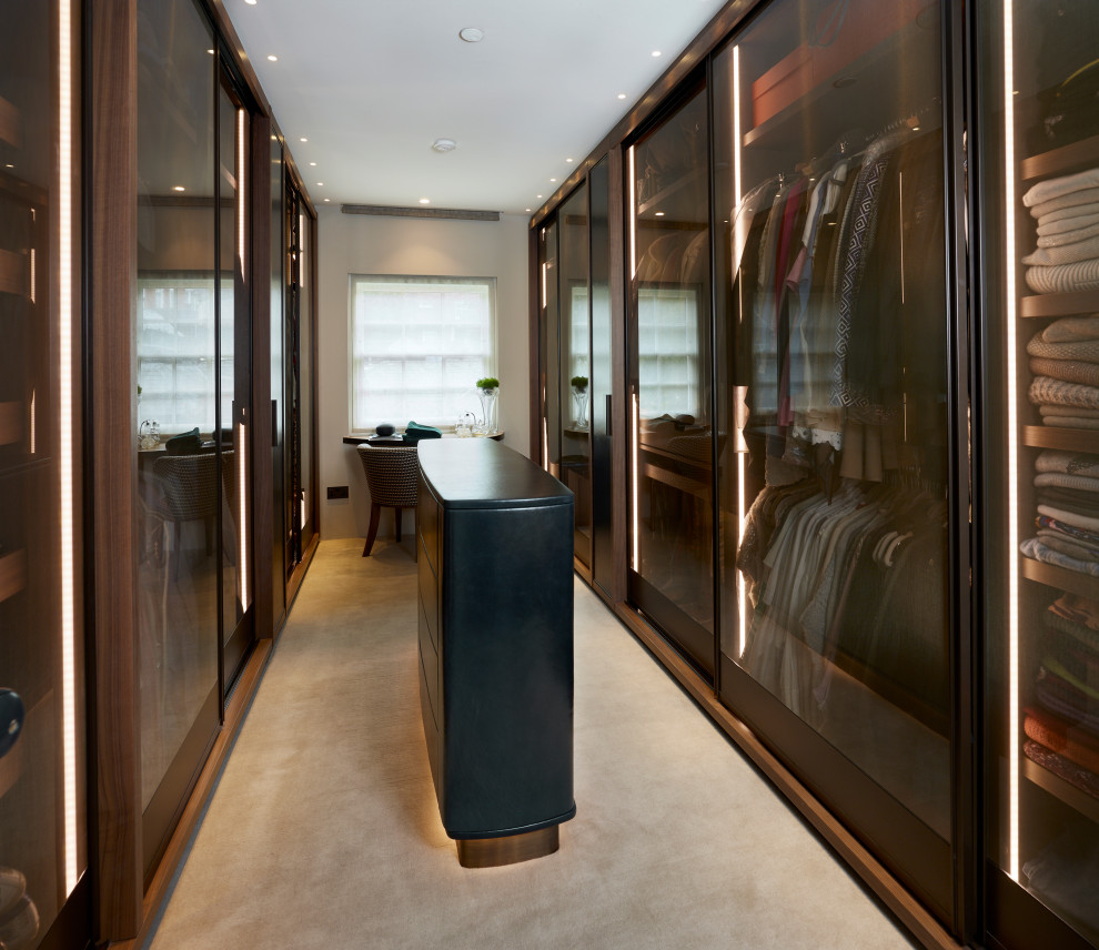 Design ideas for a large contemporary gender-neutral dressing room in London with glass-front cabinets, brown cabinets, carpet and beige floor.