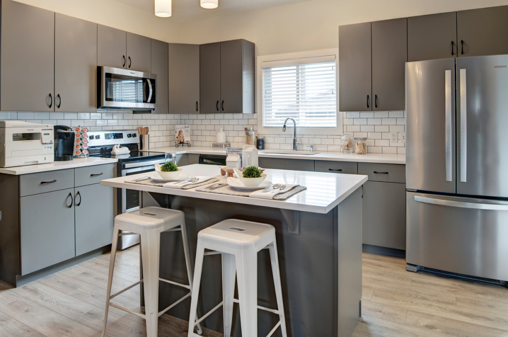 This is an example of a small transitional l-shaped eat-in kitchen in Edmonton with an undermount sink, flat-panel cabinets, grey cabinets, quartzite benchtops, white splashback, ceramic splashback, stainless steel appliances, laminate floors, with island, grey floor and white benchtop.