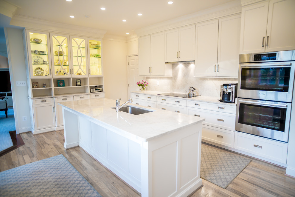 Large traditional l-shaped separate kitchen in DC Metro with a drop-in sink, glass-front cabinets, white cabinets, beige splashback, stainless steel appliances, light hardwood floors, with island, brown floor and quartzite benchtops.