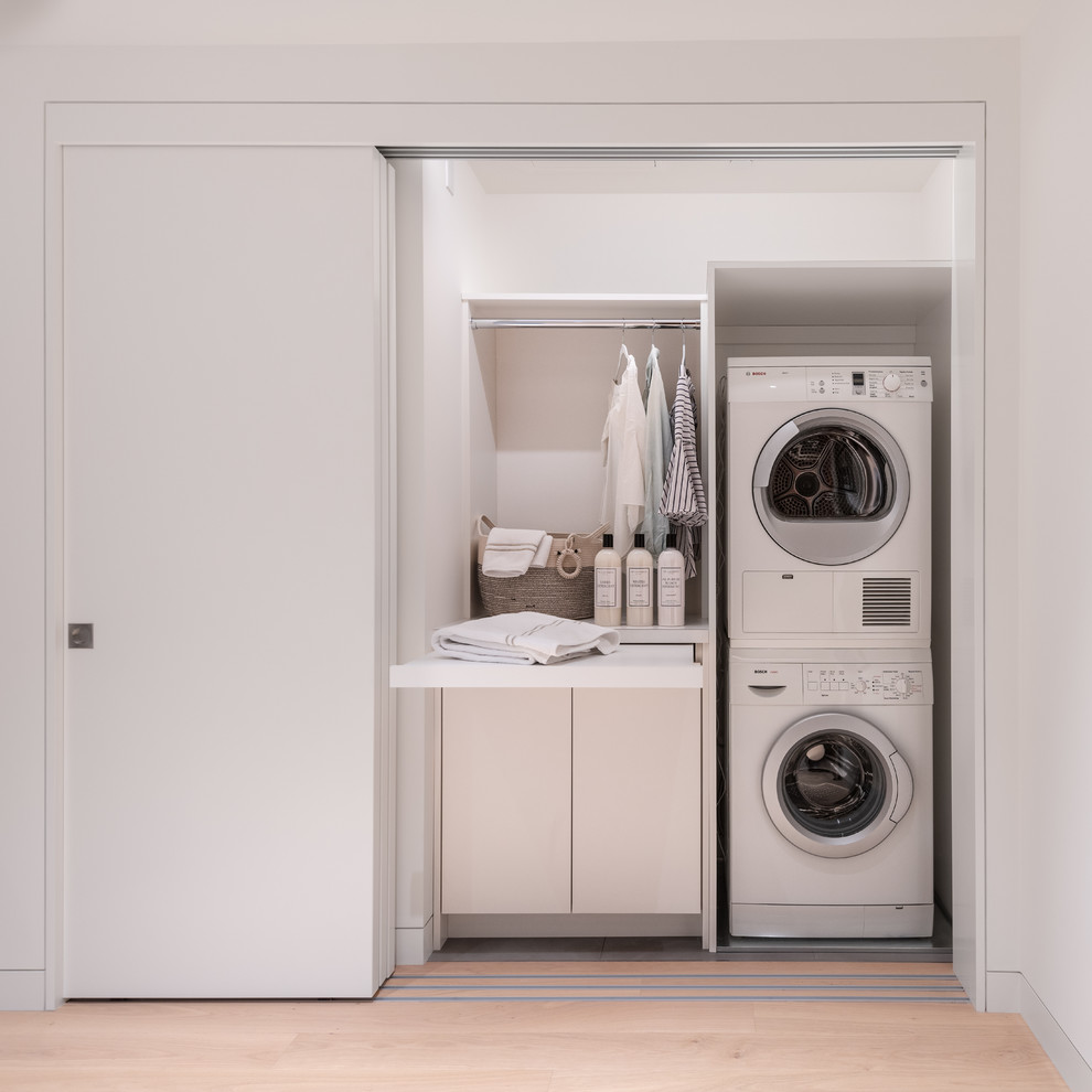 Small contemporary single-wall laundry cupboard in San Francisco with white cabinets, white walls, a stacked washer and dryer, beige floor, white benchtop, flat-panel cabinets and light hardwood floors.