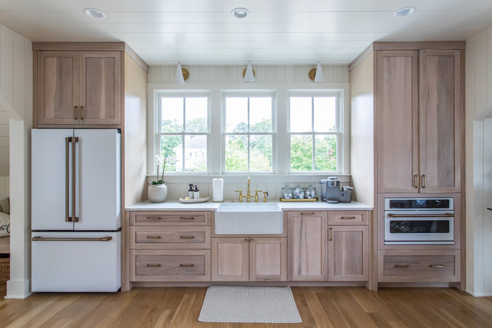 Design ideas for a transitional single-wall kitchen in Atlanta with a farmhouse sink, shaker cabinets, light wood cabinets, timber splashback, white appliances, light hardwood floors, brown floor, white benchtop and timber.