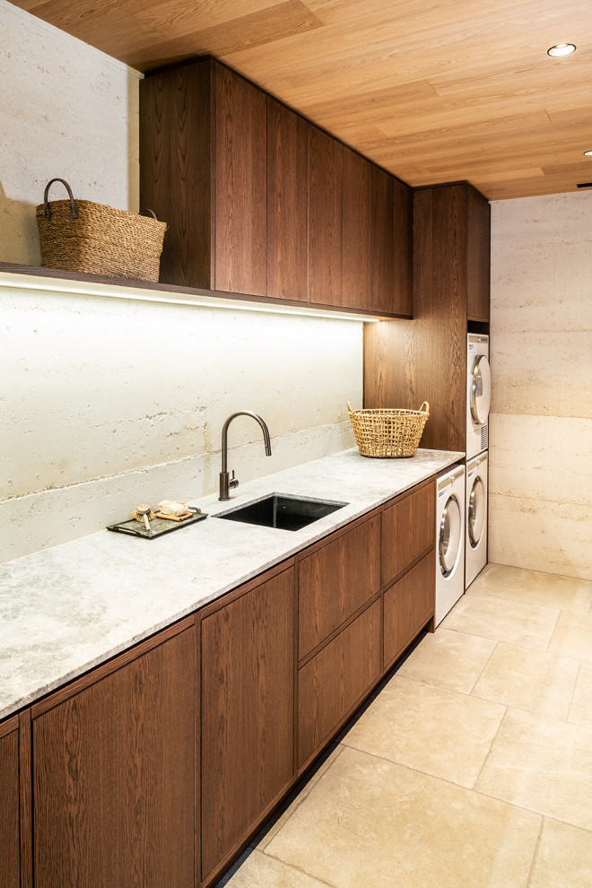 This is an example of a large contemporary galley dedicated laundry room in Melbourne with an undermount sink, flat-panel cabinets, medium wood cabinets, limestone benchtops, beige splashback, beige walls, a stacked washer and dryer, grey floor, grey benchtop and timber.