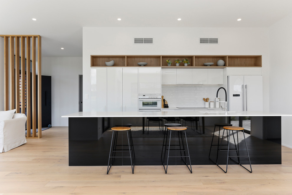 Photo of a contemporary galley kitchen in Auckland with an undermount sink, flat-panel cabinets, white cabinets, white splashback, subway tile splashback, white appliances, light hardwood floors, with island, beige floor and white benchtop.