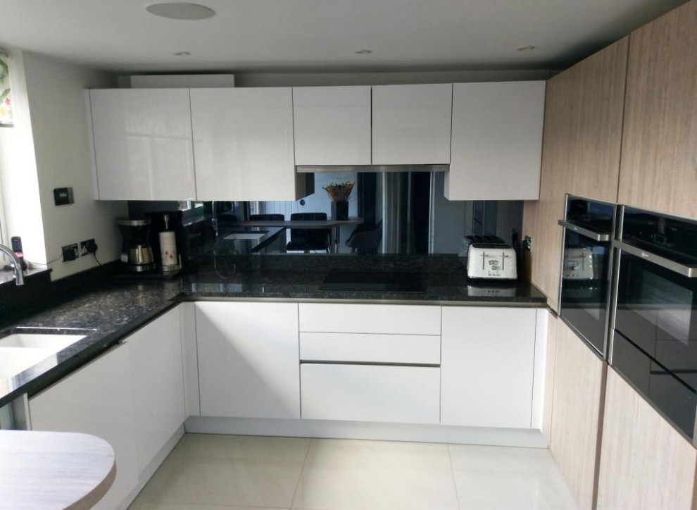 Small modern u-shaped eat-in kitchen in London with an integrated sink, flat-panel cabinets, granite benchtops, metallic splashback, glass sheet splashback, stainless steel appliances, ceramic floors, a peninsula, beige floor, black benchtop and white cabinets.