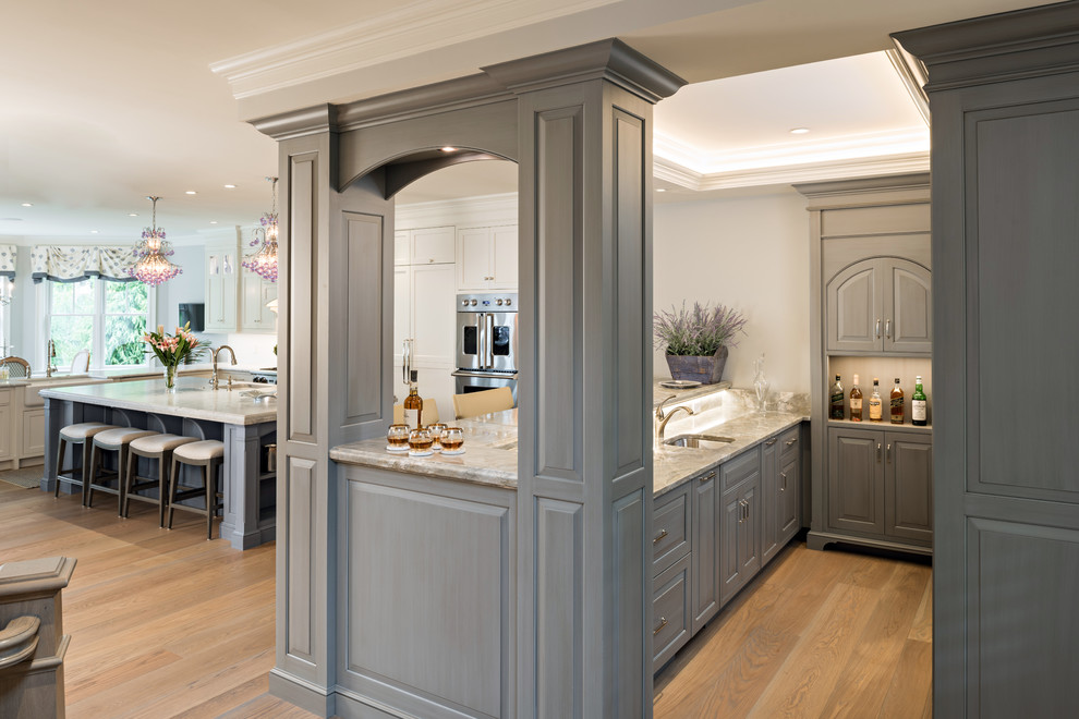 This is an example of a large eclectic l-shaped seated home bar in Boston with raised-panel cabinets, grey cabinets, marble benchtops, grey splashback, light hardwood floors, brown floor and an undermount sink.