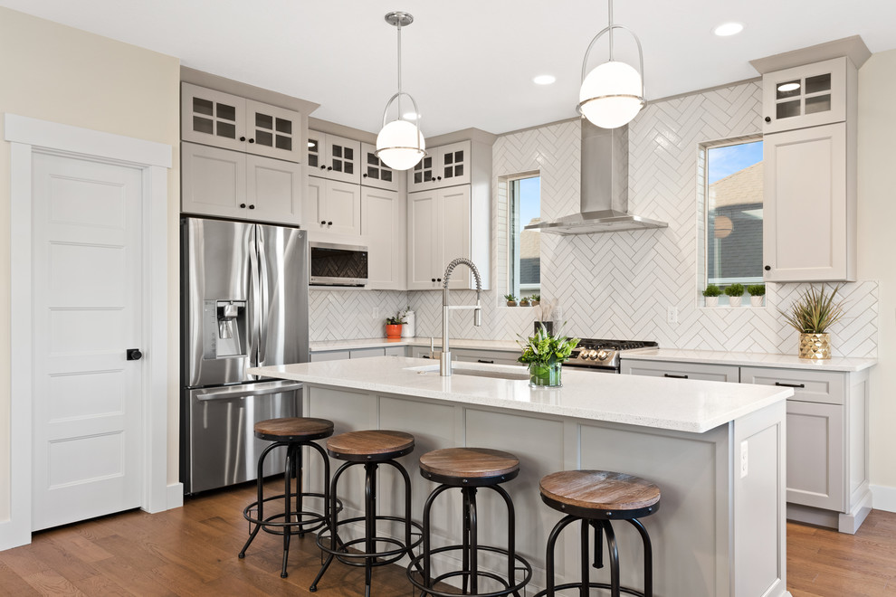 Photo of a mid-sized transitional l-shaped eat-in kitchen in Salt Lake City with a single-bowl sink, recessed-panel cabinets, grey cabinets, quartz benchtops, white splashback, porcelain splashback, stainless steel appliances, medium hardwood floors, with island and white benchtop.