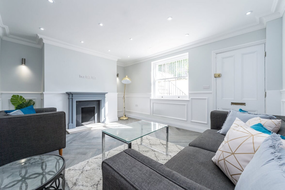 Photo of a mid-sized traditional formal open concept living room in London with grey walls, light hardwood floors, a standard fireplace, a wood fireplace surround, a wall-mounted tv and grey floor.