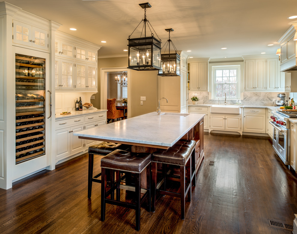 This is an example of a large traditional u-shaped open plan kitchen in Philadelphia with a farmhouse sink, raised-panel cabinets, white cabinets, marble benchtops, stone tile splashback, panelled appliances, dark hardwood floors, with island, grey splashback, brown floor and white benchtop.