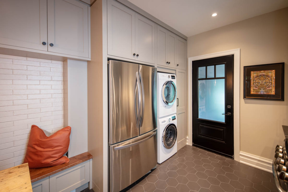 This is an example of a small country galley utility room in Ottawa with an undermount sink, shaker cabinets, medium wood cabinets, quartz benchtops, multi-coloured splashback, porcelain splashback, porcelain floors, grey floor, black benchtop, beige walls and a stacked washer and dryer.