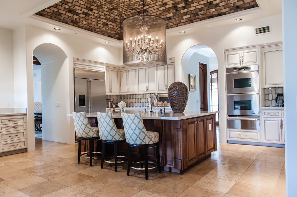 Open concept kitchen - large transitional single-wall travertine floor open concept kitchen idea in Phoenix with a farmhouse sink, raised-panel cabinets, white cabinets, marble countertops, white backsplash, stone tile backsplash, stainless steel appliances and an island