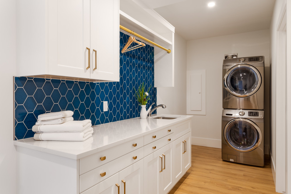 Inspiration for a mid-sized transitional galley dedicated laundry room in Calgary with a single-bowl sink, shaker cabinets, white cabinets, quartz benchtops, blue splashback, ceramic splashback, white walls, light hardwood floors, a stacked washer and dryer, beige floor and white benchtop.