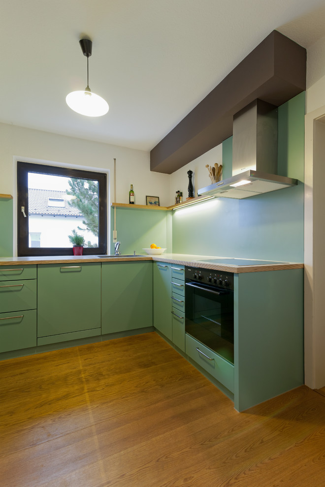 Mid-sized traditional l-shaped separate kitchen in Munich with a drop-in sink, flat-panel cabinets, green cabinets, wood benchtops, green splashback, black appliances, medium hardwood floors and no island.