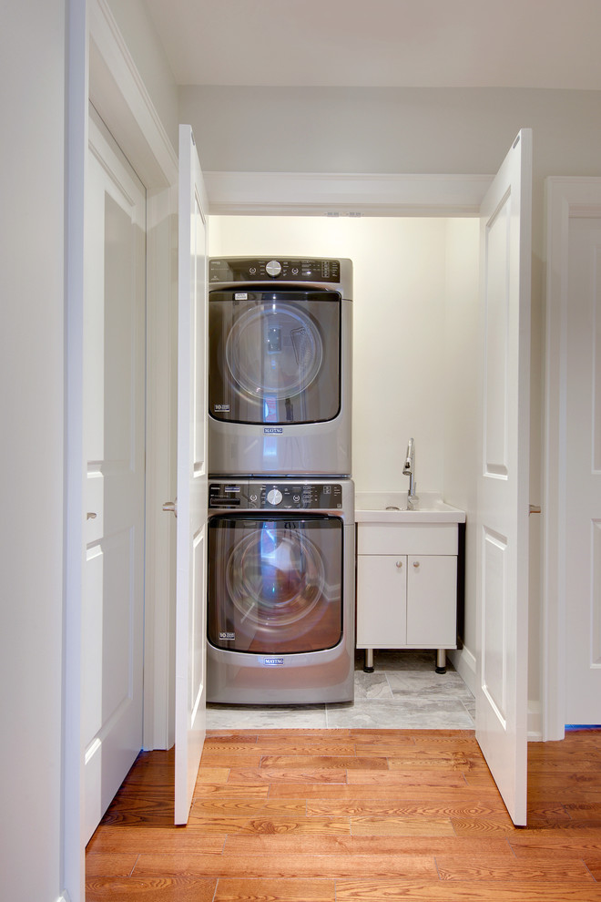 This is an example of a small transitional single-wall laundry cupboard in Toronto with an integrated sink, flat-panel cabinets, white cabinets, white walls, ceramic floors and a stacked washer and dryer.