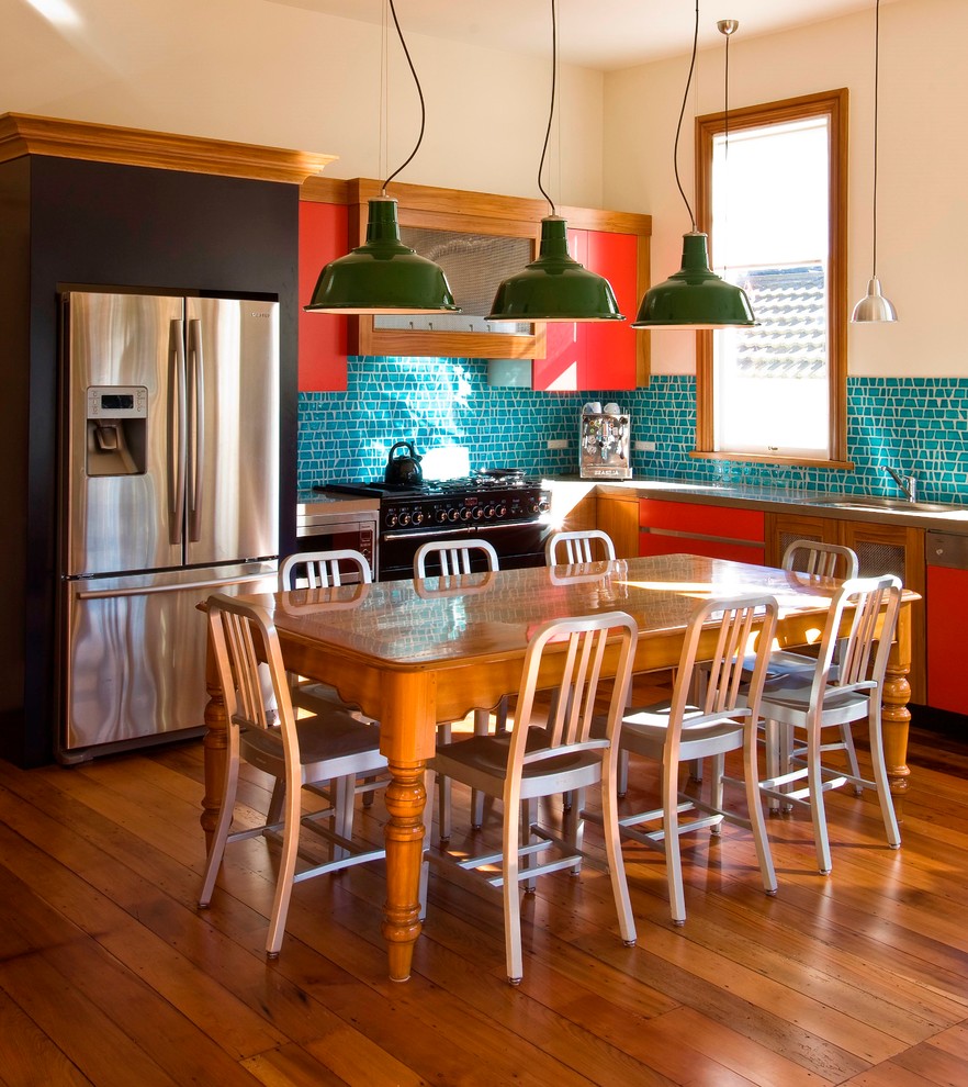 This is an example of an eclectic l-shaped eat-in kitchen in Auckland with flat-panel cabinets, red cabinets, mosaic tile splashback and stainless steel appliances.
