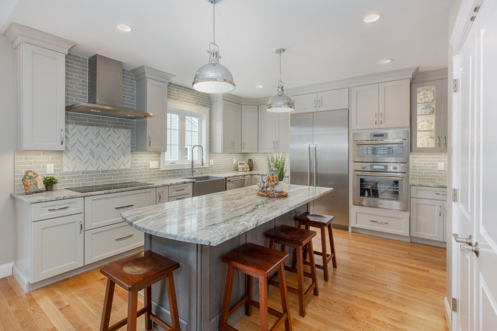 This is an example of a large transitional l-shaped eat-in kitchen in Boston with a farmhouse sink, grey cabinets, grey splashback, stainless steel appliances, light hardwood floors, with island, brown floor, shaker cabinets and grey benchtop.