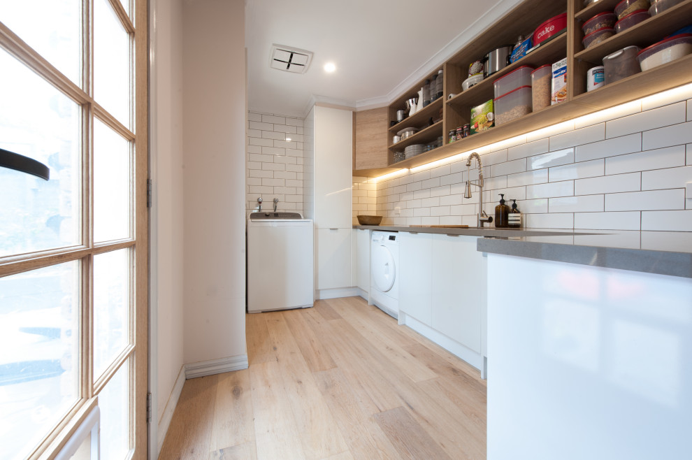 Design ideas for a mid-sized scandinavian galley utility room in Melbourne with a double-bowl sink, open cabinets, white cabinets, solid surface benchtops, white splashback, subway tile splashback, white walls, light hardwood floors, an integrated washer and dryer and grey benchtop.