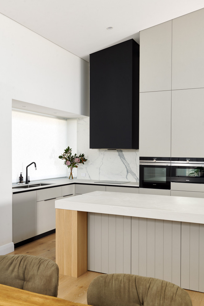 Inspiration for a large contemporary l-shaped kitchen/diner in Perth with a double-bowl sink, flat-panel cabinets, grey cabinets, engineered stone countertops, white splashback, porcelain splashback, black appliances, light hardwood flooring, an island, beige floors, white worktops and a vaulted ceiling.