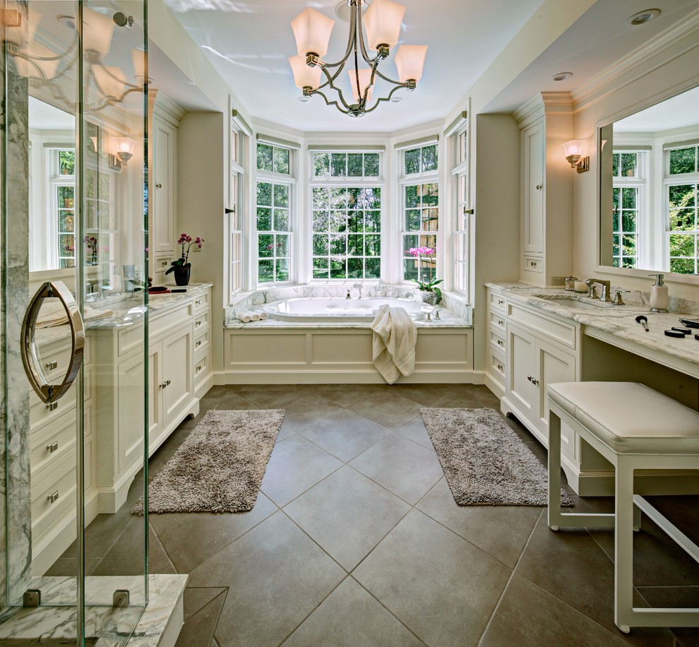 This is an example of a large traditional master bathroom in New York with an undermount sink, recessed-panel cabinets, white cabinets, a drop-in tub, beige walls, marble benchtops, a corner shower, a hinged shower door, white tile, marble and grey floor.