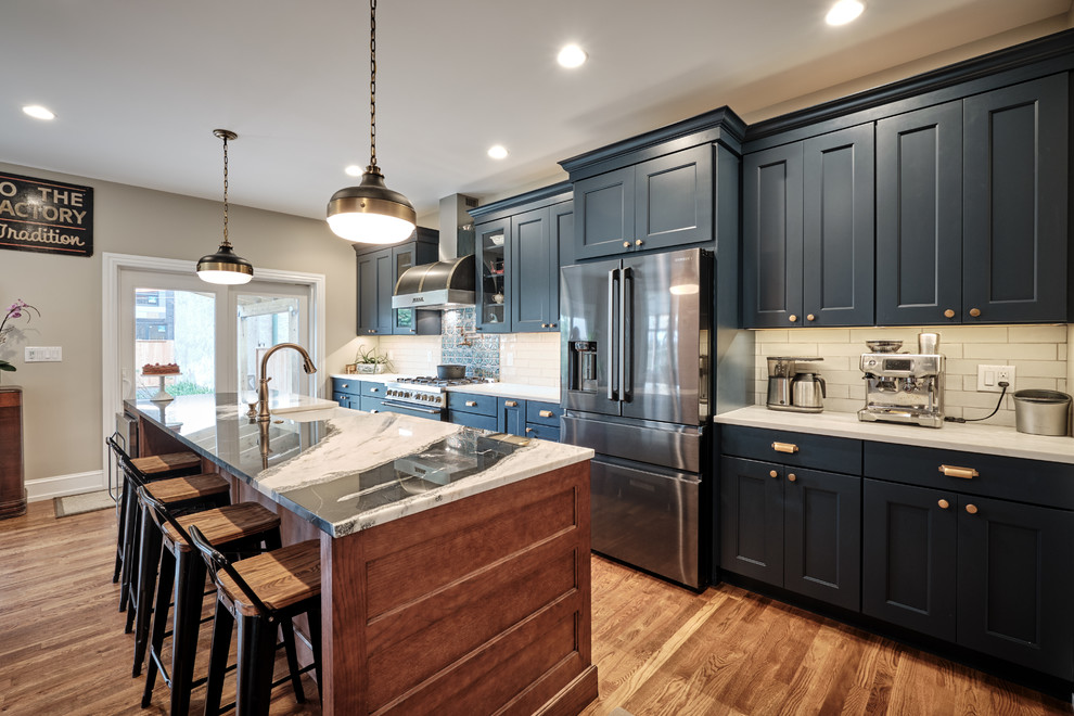 Large eclectic galley open plan kitchen in Philadelphia with a farmhouse sink, recessed-panel cabinets, blue cabinets, quartzite benchtops, grey splashback, ceramic splashback, black appliances, medium hardwood floors, with island, brown floor and multi-coloured benchtop.