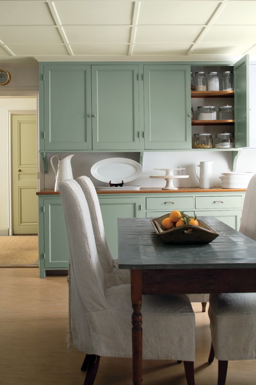 Sage-green Shaker kitchen cabinets with wood countertop and open shelves; rustic wood dining table and slipcovered chairs in a bright, cozy space