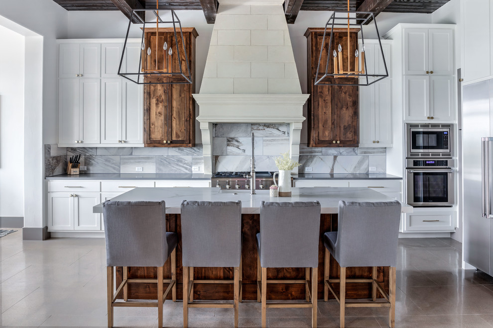 Mid-sized transitional l-shaped open plan kitchen in Austin with shaker cabinets, white cabinets, white splashback, stainless steel appliances, with island, grey floor, grey benchtop, a farmhouse sink, quartz benchtops, marble splashback and cement tiles.