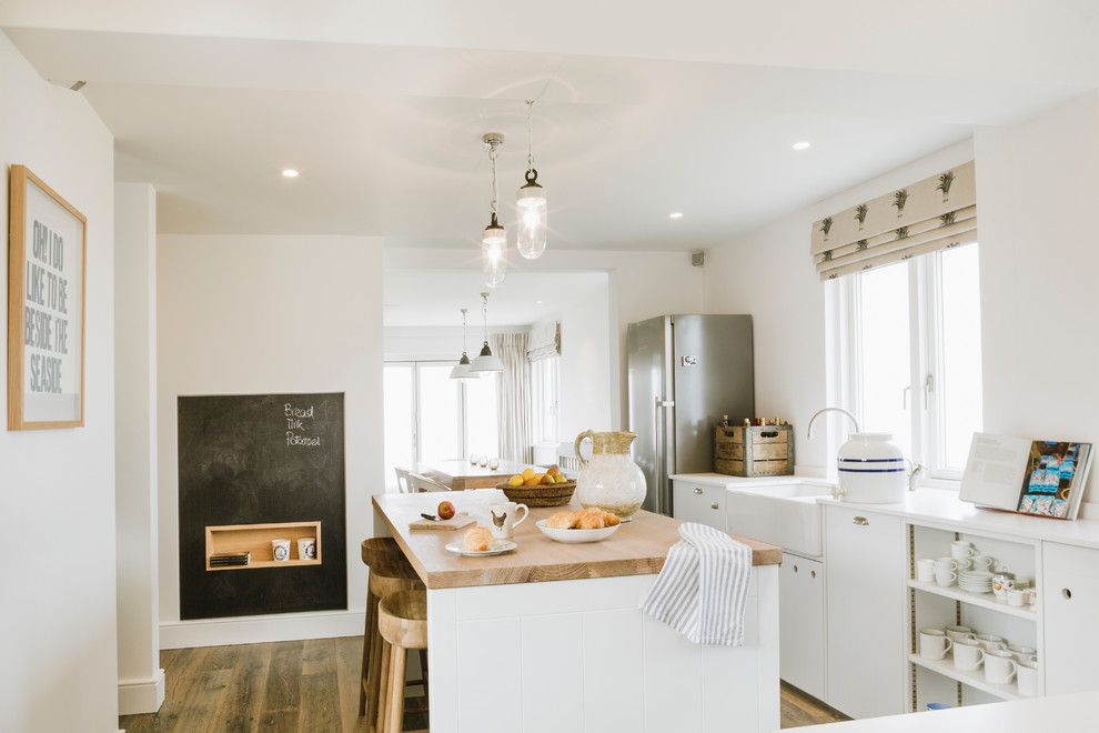 Mid-sized beach style eat-in kitchen in Hampshire with a farmhouse sink, flat-panel cabinets, white cabinets, stainless steel appliances, medium hardwood floors, with island and wood benchtops.