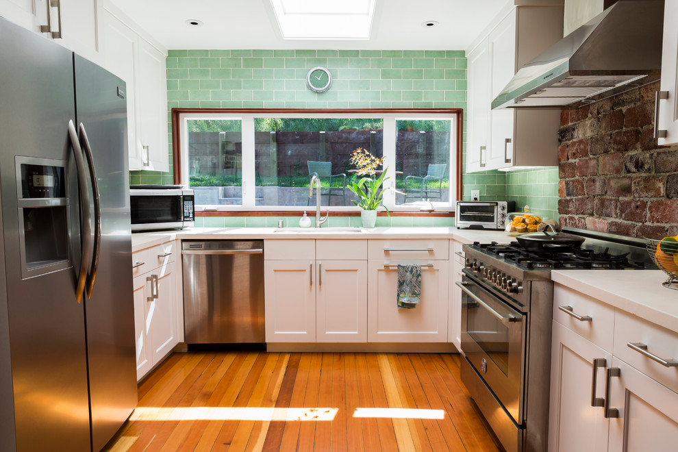 This is an example of a mid-sized transitional u-shaped open plan kitchen in San Francisco with an undermount sink, shaker cabinets, white cabinets, limestone benchtops, green splashback, glass tile splashback, stainless steel appliances, light hardwood floors and no island.