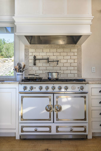 Large country open plan kitchen in San Francisco with a farmhouse sink, white cabinets, limestone benchtops, beige splashback, subway tile splashback, white appliances, light hardwood floors and with island.