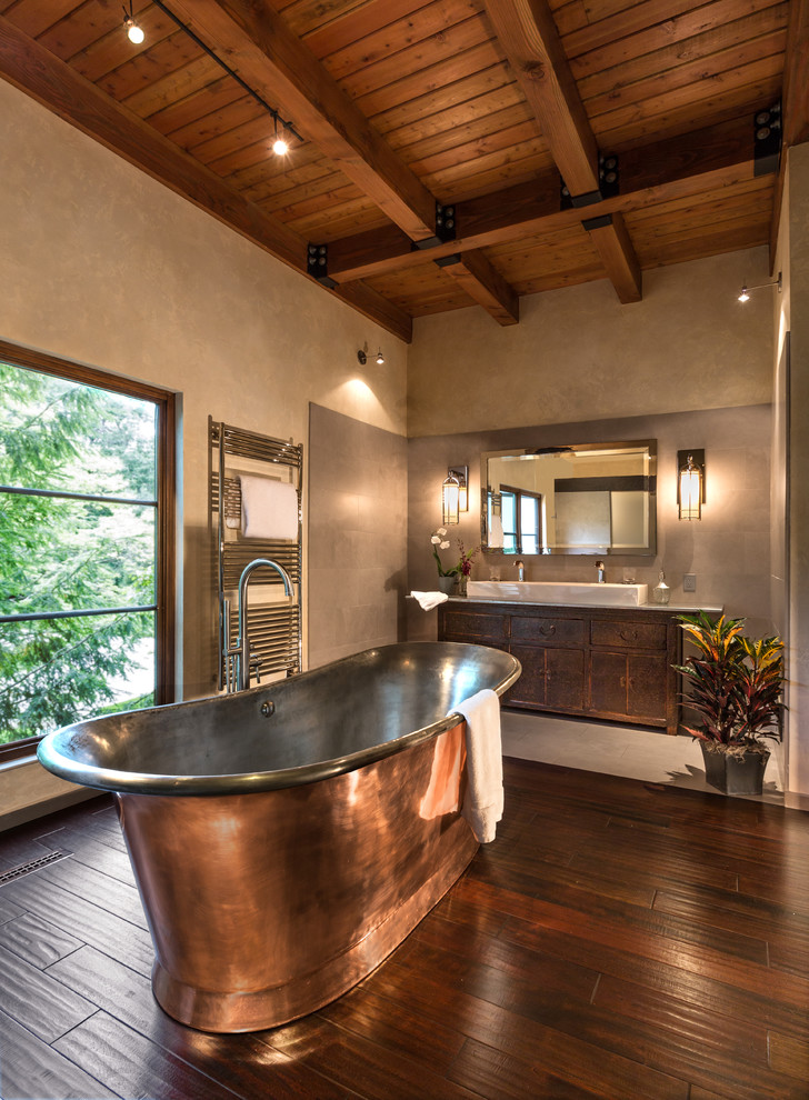Photo of a country master bathroom in Milwaukee with recessed-panel cabinets, a freestanding tub, an alcove shower, a one-piece toilet, gray tile, stone tile, beige walls, dark hardwood floors, a trough sink and dark wood cabinets.