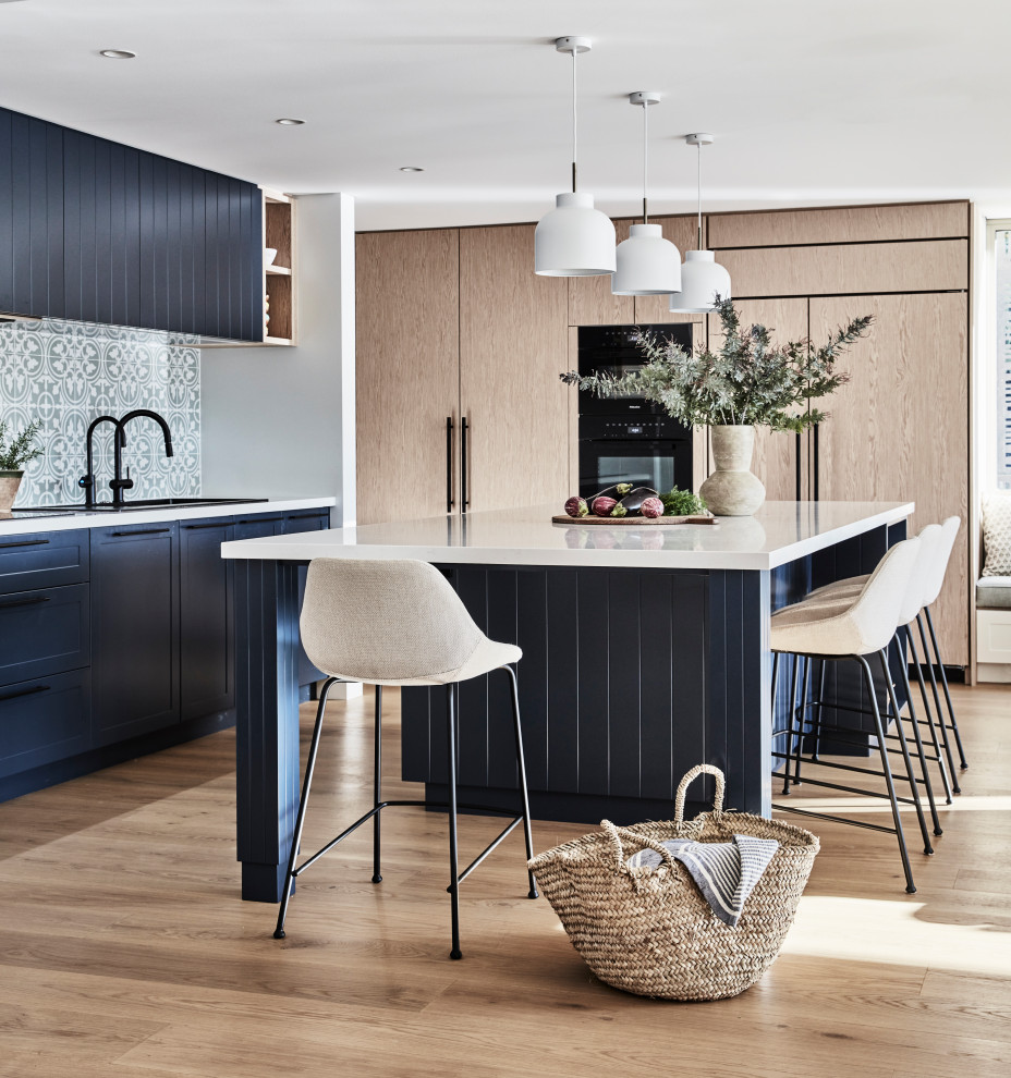 Inspiration for an expansive beach style l-shaped open plan kitchen in Sydney with a built-in sink, shaker cabinets, blue cabinets, engineered stone countertops, green splashback, porcelain splashback, black appliances, light hardwood flooring, an island, beige floors and white worktops.