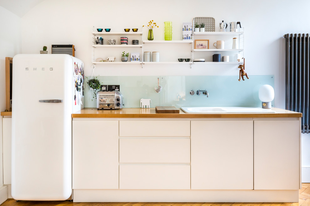 This is an example of a scandinavian single-wall kitchen in London with a drop-in sink, flat-panel cabinets, white cabinets, wood benchtops, glass sheet splashback and medium hardwood floors.