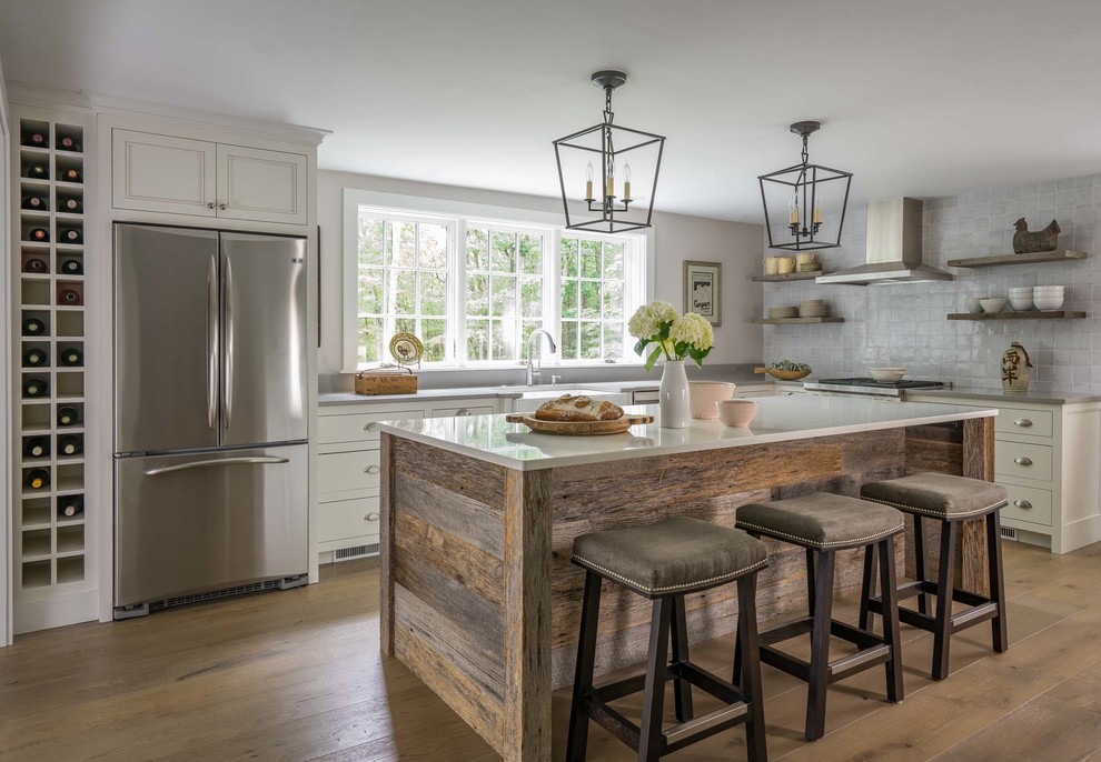 This is an example of a mid-sized country l-shaped kitchen in Boston with a farmhouse sink, recessed-panel cabinets, white cabinets, quartzite benchtops, grey splashback, porcelain splashback, stainless steel appliances, light hardwood floors, with island and brown floor.