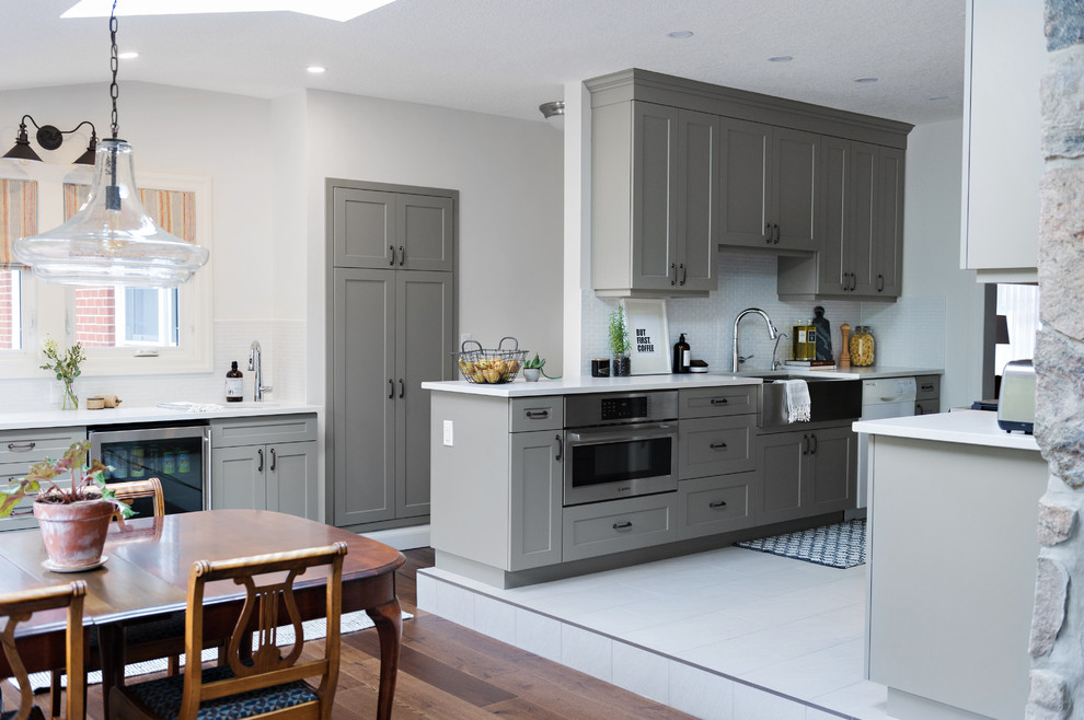 Inspiration for a large transitional l-shaped dark wood floor and brown floor enclosed kitchen remodel in Toronto with shaker cabinets, gray cabinets, no island, a farmhouse sink, quartz countertops, white backsplash, porcelain backsplash and stainless steel appliances