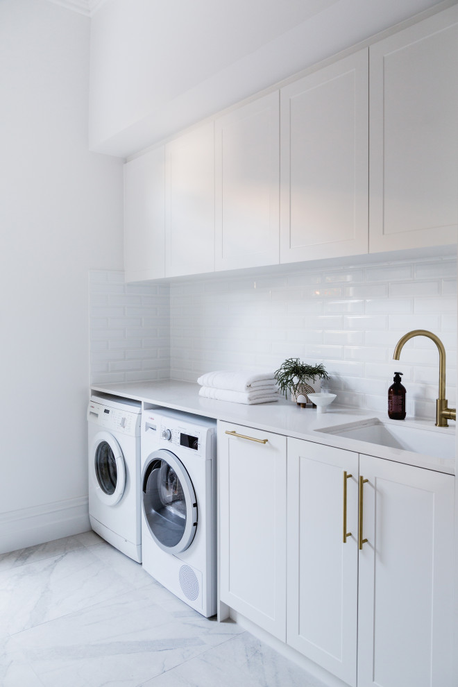 Photo of a mid-sized traditional single-wall dedicated laundry room in Melbourne with an undermount sink, shaker cabinets, white cabinets, limestone benchtops, white splashback, ceramic splashback, white walls, painted wood floors, a side-by-side washer and dryer, brown floor and white benchtop.