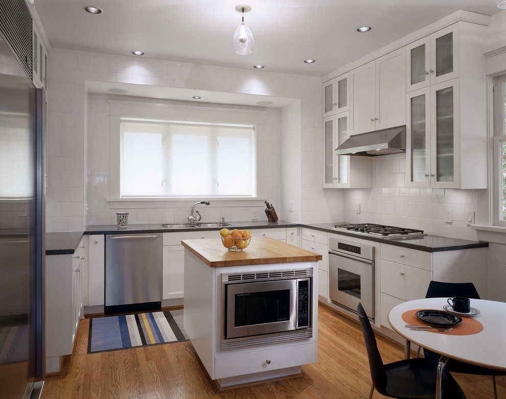 This is an example of a traditional u-shaped eat-in kitchen in Seattle with stainless steel appliances, wood benchtops, white splashback, an undermount sink, glass-front cabinets, white cabinets and subway tile splashback.