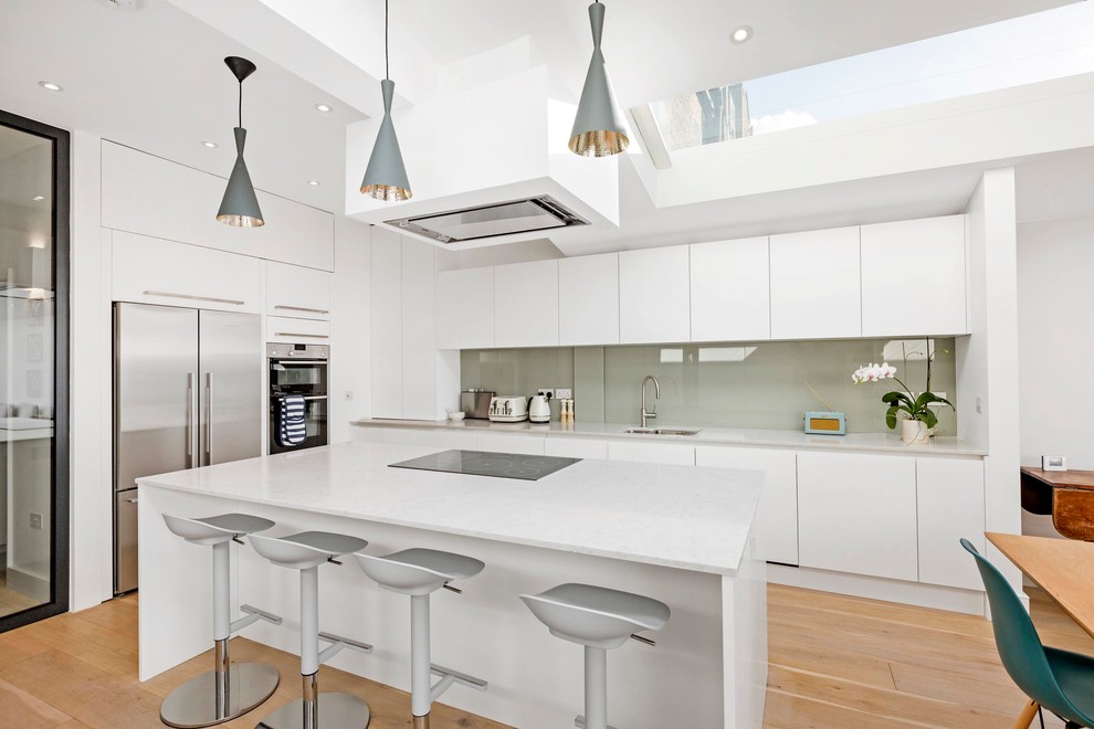 This is an example of a contemporary l-shaped open plan kitchen in London with an undermount sink, flat-panel cabinets, white cabinets, grey splashback, glass sheet splashback, stainless steel appliances, light hardwood floors, with island and brown floor.