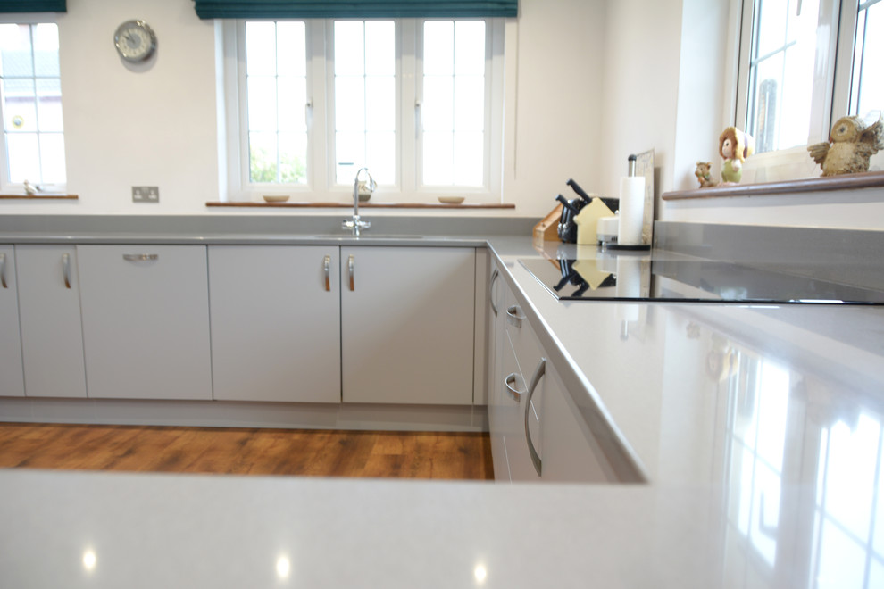 This is an example of a mid-sized contemporary u-shaped eat-in kitchen in Cornwall with a drop-in sink, flat-panel cabinets, grey cabinets, solid surface benchtops, grey splashback, black appliances, medium hardwood floors, a peninsula and grey benchtop.